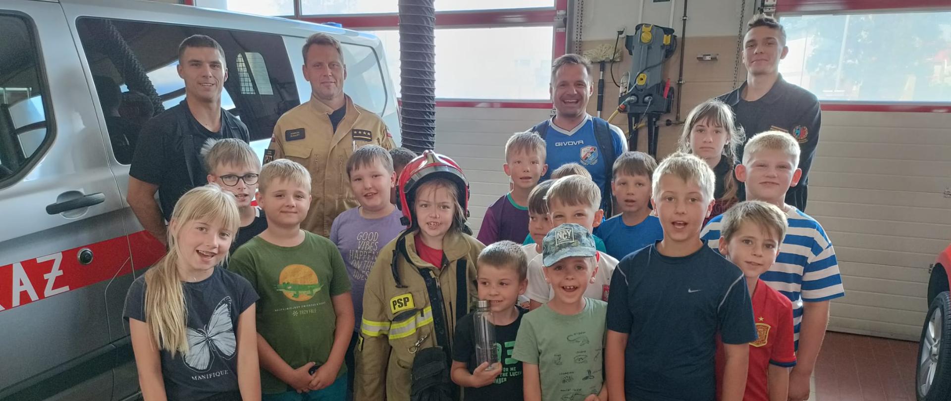 Zdjęcie przedstawia dzieci wraz ze strażakami pozującymi do zdjęcia w garażu strażnicy.