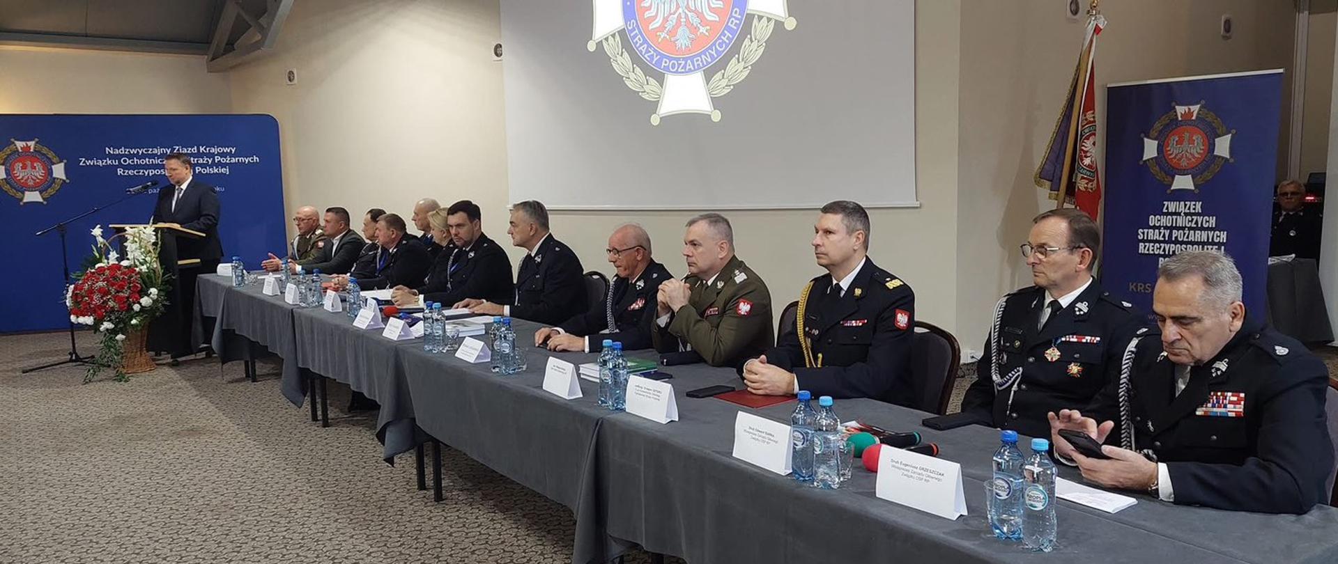 Wewnątrz sali konferencyjnej przy stołach znajdują się strażacy ochotnicy ubrani w mundury wyjściowe. Wszyscy siedzą przy stołach. Na rzutnikach wyświetlone logo związku OSP. W centrum zdjęcia przy mównicy przemawia Marcin Kierwiński - minister spraw wewnętrznych i administracji. 