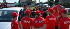 Obóz survivalowy w Sobiałkowie. Zdjęcie przedstawia grupę dziewcząt i chłopców stojących obok samochodu. Częśc z nich ma na sobie czerwone czapki oraz koszulki z białym napisem "STRAŻ", pozostali ubrani są w koszulki i czapki koloru czarnego. Obok samochodu stoi również strażak PSAP, który dystykulując objaśnia słuchaczom w jaki sposób ewakuować poszkodowanego z samochodu. W tle namiot i wóz strażacki.
