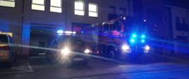  Dwa samochody pożarnicze stojak przed budynkiem remizy z włączonymi światłami błyskowymi z boku widać kawałek srebrnego samochodu