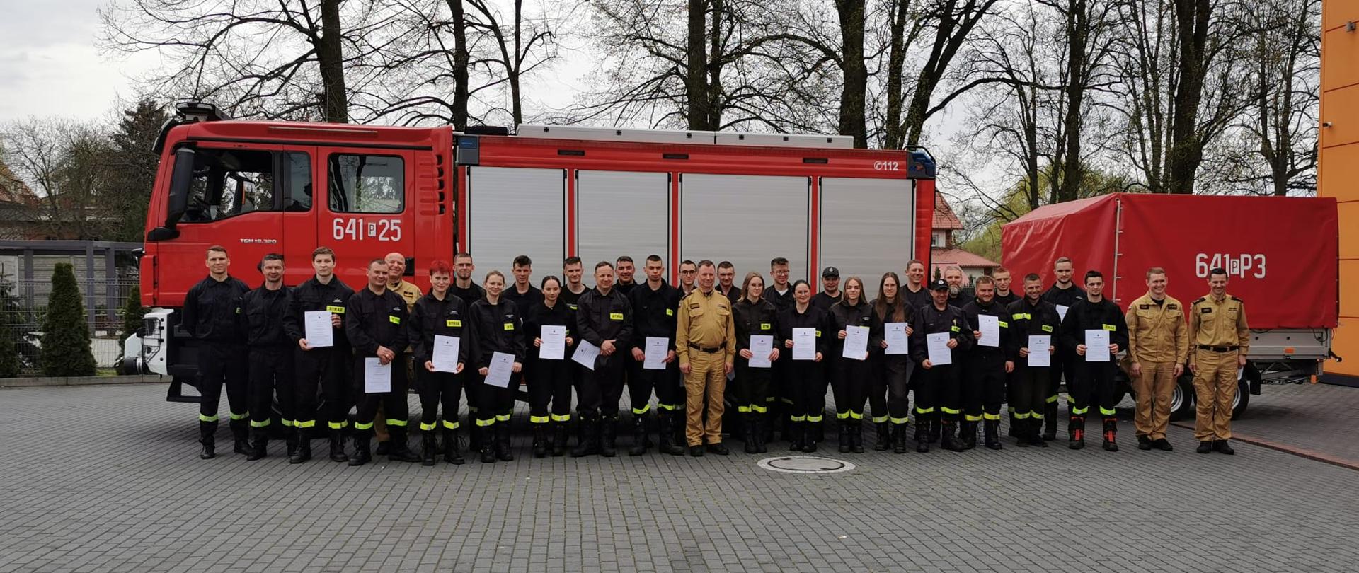 Zdjęcie grupowe absolwentów szkolenia oraz funkcjonariuszy PSP na tle samochodu strażackiego.