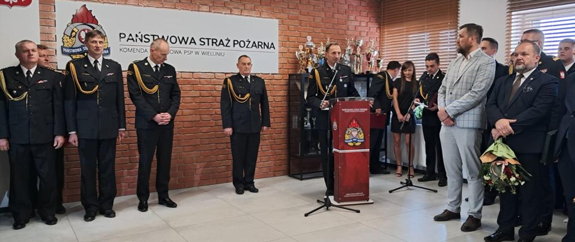 Grupa strażaków w mundurach stoi w pomieszczeniu biurowym przed ścianą z czerwonej cegły i tablicą z napisem "PAŃSTWOWA STRAŻ POŻARNA". Jeden z mężczyzn w mundurze przemawia z mównicy. Po prawej stronie widać cywilów, z których jeden trzyma bukiet kwiatów. Na tylnym planie widać regał z pucharami.