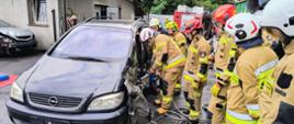 Kolorowa fotografia wykonana na zewnątrz, przedstawia strażaków ochotniczych straży pożarnych stojących w ubraniach specjalnych i hełmach podczas szkolenia praktycznego z zakresu ratownictwa technicznego. Strażacy wykonują dostęp do poszkodowanego kierowcy.