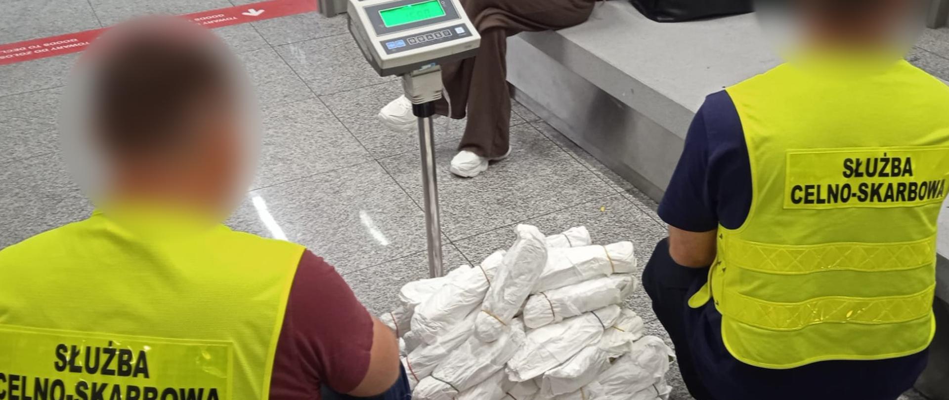 Dwie osoby w kamizelkach „Służba Celno‑Skarbowa” przy wadze elektronicznej; na platformie ułożono liczne białe pakunki owinięte papierem.