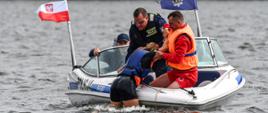 Policjanci oraz ratownik WOPR płynący łodzią policyjną wyciągają z wody kobietę. Ratownik WOPR ubrany w pomarańczową kamizelkę ratunkową. Łódź posiada dwie flagi: Polską oraz formacji Policji. 