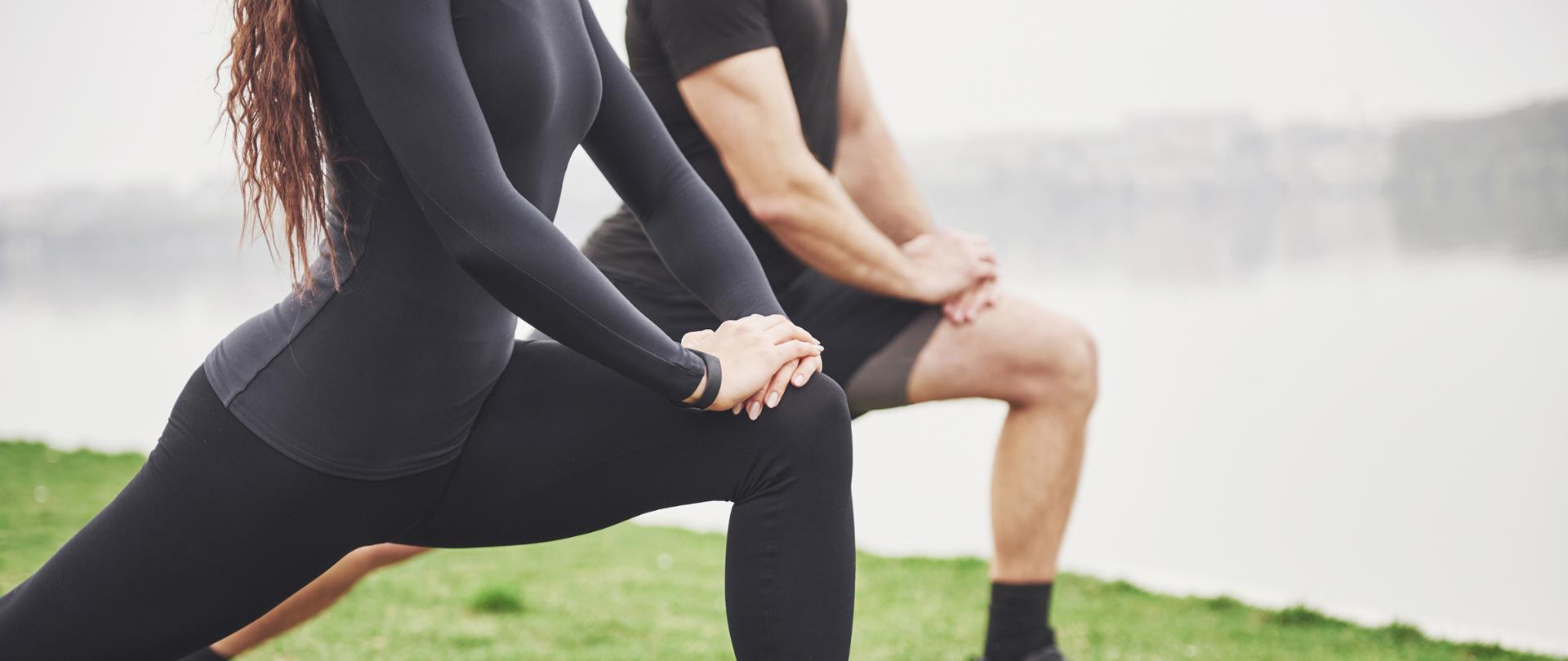Fitness couple stretching outdoors in park near the water. Young bearded man and woman exercising together in morning.