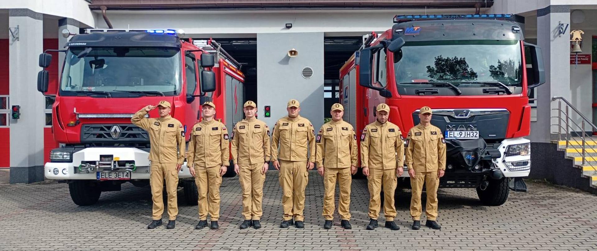 Na zdjęciu widzimy siedmiu strażaków ubranych w mundury koloru piaskowego, stojących w szeregu przed dwoma czerwonymi wozami strażackimi. Pojazdy te zaparkowane są przed budynkiem straży pożarnej. Budynek ma biało-szarą elewację z czerwonymi drzwiami garażowymi oraz oknami na piętrze. 