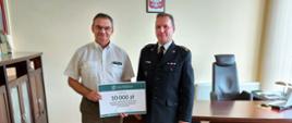 Zdjęcie przedstawia Nadleśniczego w mundurze lasów państwowych oraz komendanta powiatowego w mundurze wyjściowym. Panowie stoją na tle jasnej ściany i przekazują sobie symboliczny czek wartości 10.000 zł. Na ścianie jest zawieszone godło Polski. 