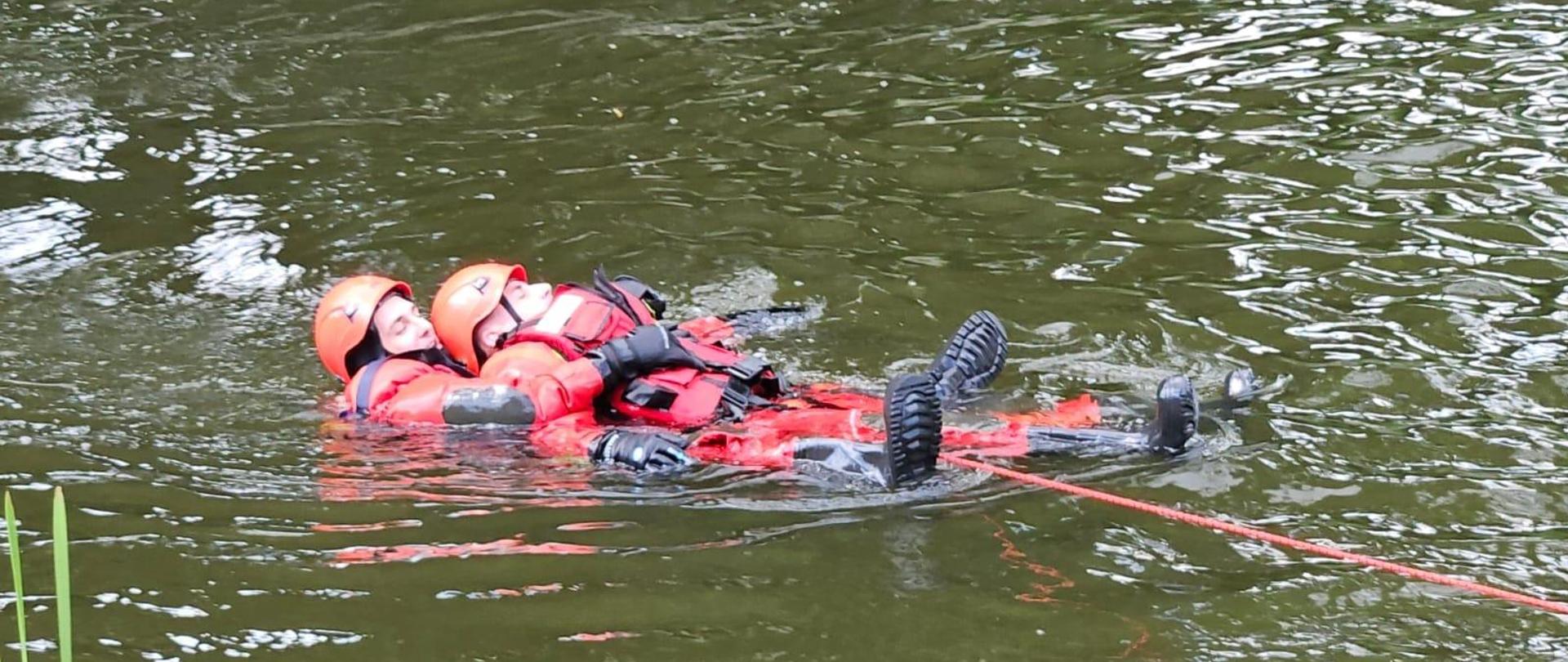 Dwóch strażaków w czerwonych kombinezonach i kaskach unosi się na wodzie.