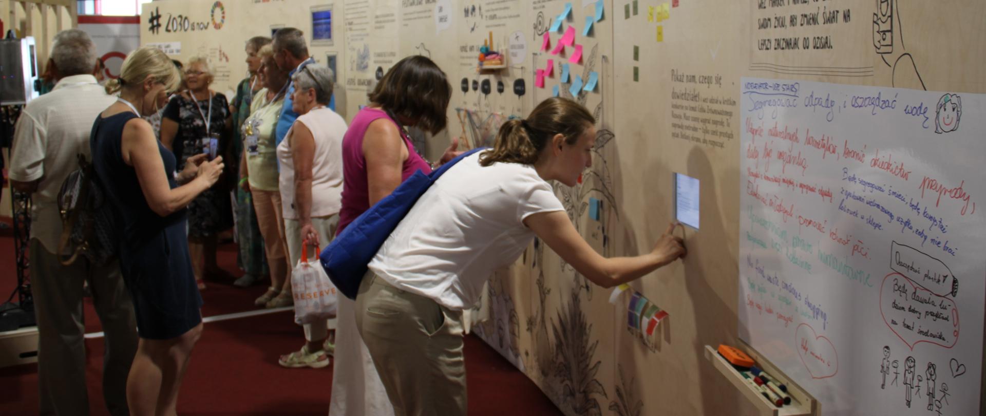 Kolorowa ścianka edukacyjna Polskiej pomocy i Komisji Europejskiej DG DEVCO z grupą ludzi czytających informacje znajdujące się na niej i fotografujących się na jej tle