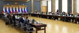 Szeroki kadr. Uczestnicy podczas posiedzenia, siedzą przy stołach konferencyjnych ustawionych w kształt litery U. U szczytu stołu między innymi Sekretarz Stanu Adriana Porowska. W tle flagi Polski i Unii Europejskiej. 