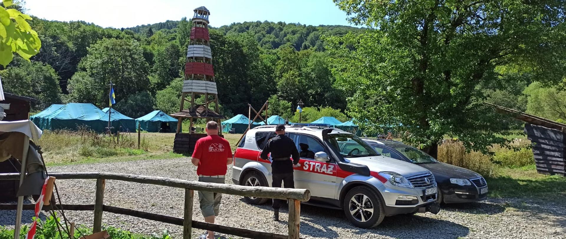 Na zdjęciu widać samochód Państwowej Straży Pożarnej przy którym stoi funkcjonariusz w ubraniu służbowym obok przechodzi Pan z obozu harcerskiego. W oddali widać wieżę widokową oraz namioty harcerzy. 