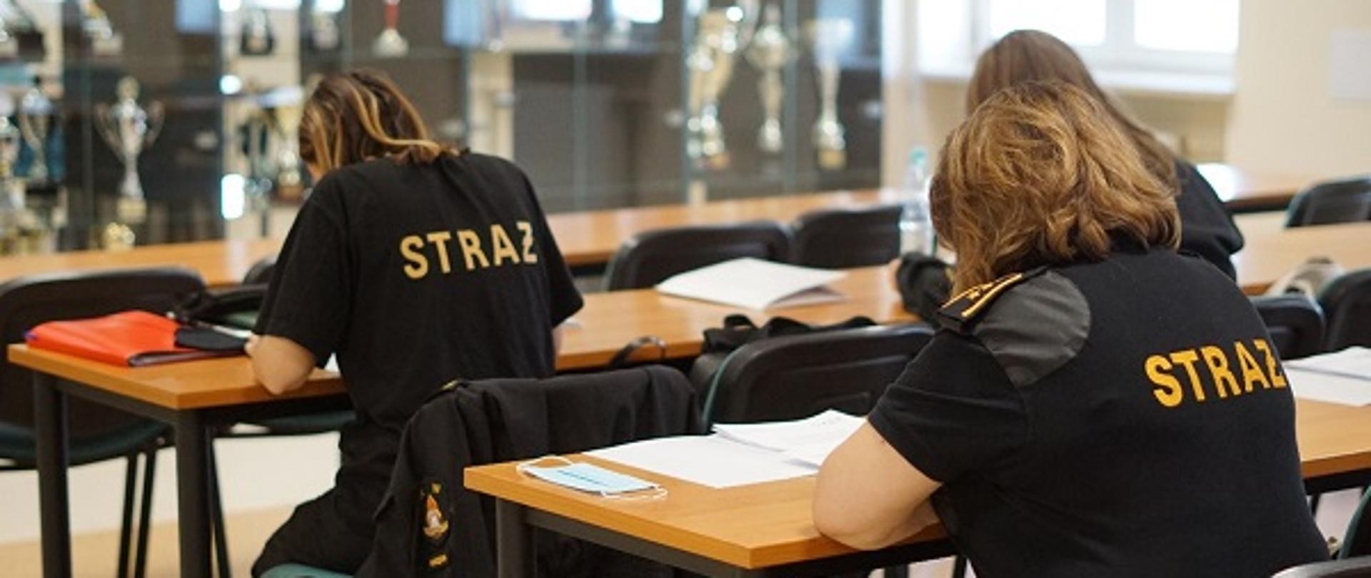 Zdjęcie przedstawia kobiety siedzące przy biurkach, na których znajdują się materiały szkoleniowe. Kobiety siedzą tyłem do zdjęcia i na czarnych koszulkach mają na plecach żółty napis Straż.
