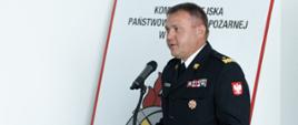 Zdjęcie zrobione w pomieszczeniu. Przy mównicy stoi oficer Państwowej Straży Pożarnej w mundurze wyjściowy, w stopniu generała. Strażak przemawia do mikrofonu. Na mównicy umieszczony jest napis: Komenda Miejska Państwowej Straży Pożarnej w Rzeszowie.