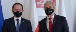 Na zdjęciu dwóch mężczyzn, stojących na tle obrazu z godłem Polski oraz flagi Unii Europejskiej i NATO. Po lewej stronie stoi konsul Węgier - młody mężczyzna, ubrany w ciemny garnitur. Na twarzy ma maseczkę ochronną z flagą Węgier. Po prawej stronie kadru, w bliskiej odległości od konsula, stoi wojewoda opolski. On również ubrany jest w ciemny garnitur, na twarzy ma czarną maseczkę z napisem Wojewoda Opolski. Przed mężczyznami, w dolnej części kadru widać fragment biurka, na którym postawiono flagietki: węgierską i polską. 