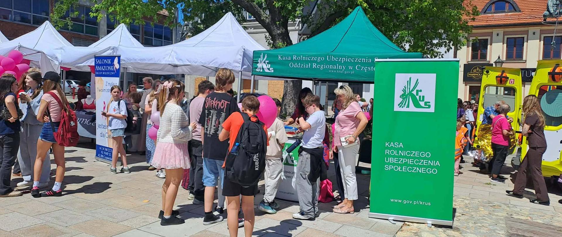 Grupa kobiet i dzieci na placu miasta przy stoisku informacyjnym KRUS. Przy stoisku pod zielonym namiotem z logo KRUS stoi kobieta w różowej bluzce i rozmawia z dziećmi, które odwiedziły stoisko. Obok zielony rollup KRUS.