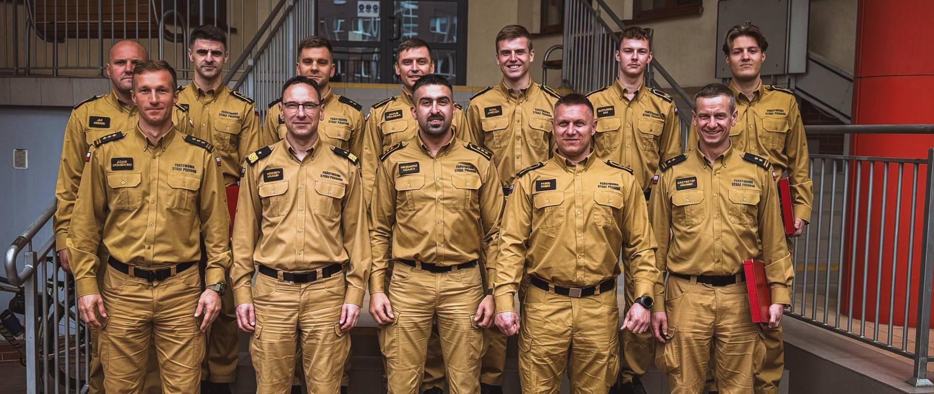 Pamiątkowe zdjęcie grupowe kadry kierowniczej oraz strażaków Państwowej Straży Pożarnej w piaskowych mundurach, pozujących na schodach przed budynkiem instytucji.