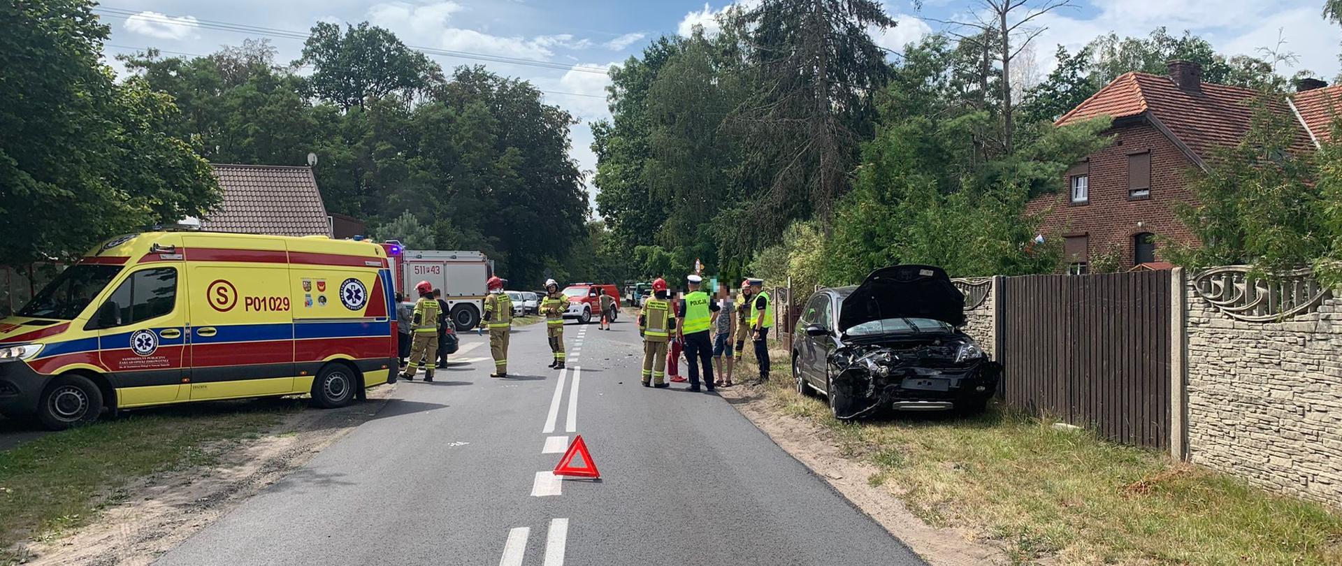 Pas drogi, na jezdni trójkąt ostrzegawczy, po prawej stronie samochód osobowy z uszkodzonym przodem, w tle strażacy i policjanci.