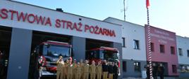 Grupa strażaków stojących przed budynkiem straży pożarnej. Na budynku widnieje napis "PAŃSTWOWA STRAŻ POŻARNA". Przed strażakami stoi jeden strażak w mundurze, który salutuje. W tle widać dwa czerwone wozy strażackie. Po prawej stronie zdjęcia znajduje się maszt z flagą Polski oraz kilku strażaków salutujących. Na budynku po prawej stronie widnieje napis "KOMENDA MIEJSKA PAŃSTWOWEJ STRAŻY POŻARNEJ W ŻORACH".