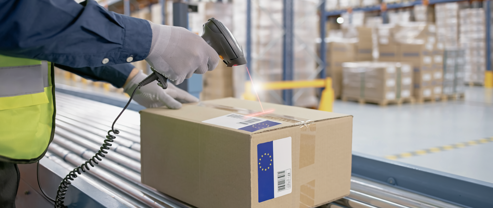 Pracownik magazynu w kamizelce odblaskowej skanuje kod kreskowy na kartonowej paczce znajdującej się na taśmie transportowej; w tle widoczne regały z towarem.