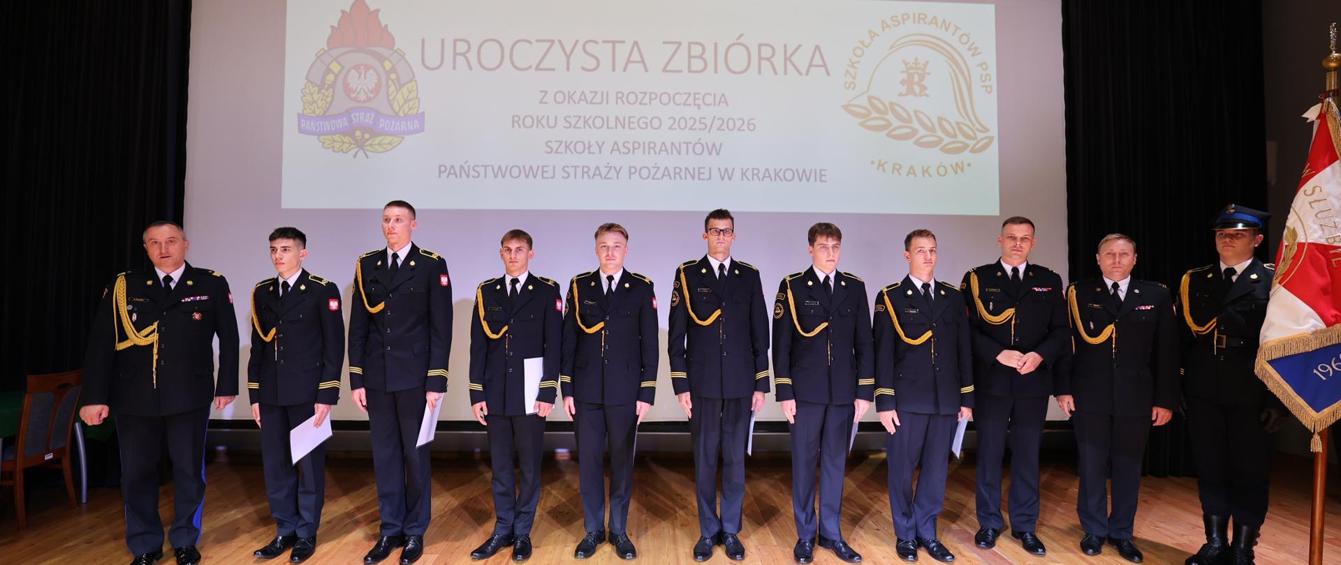 Scena auli Szkoły Aspirantów PSP w Krakwie – zdjęcie pamiątkowe uczestników uroczystej zbiórki. Na tle białego ekranu stają ubrani w wyjściowe mundury strażacy. W rękach trzymają białe teczki z otrzymanymi dokumentami o awansie lub wyróżnieniu.