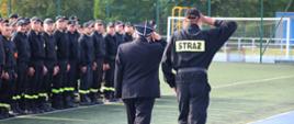Prezes Zarządu Oddziału Powiatowego ZOSP RP w Mińsku Mazowieckim wraz z dowódcą pododdziału dokonują przeglądu pododdziału. Idą po bieżni wzdłuż strażaków stojących na murawie w dwuszeregu i oddają honor