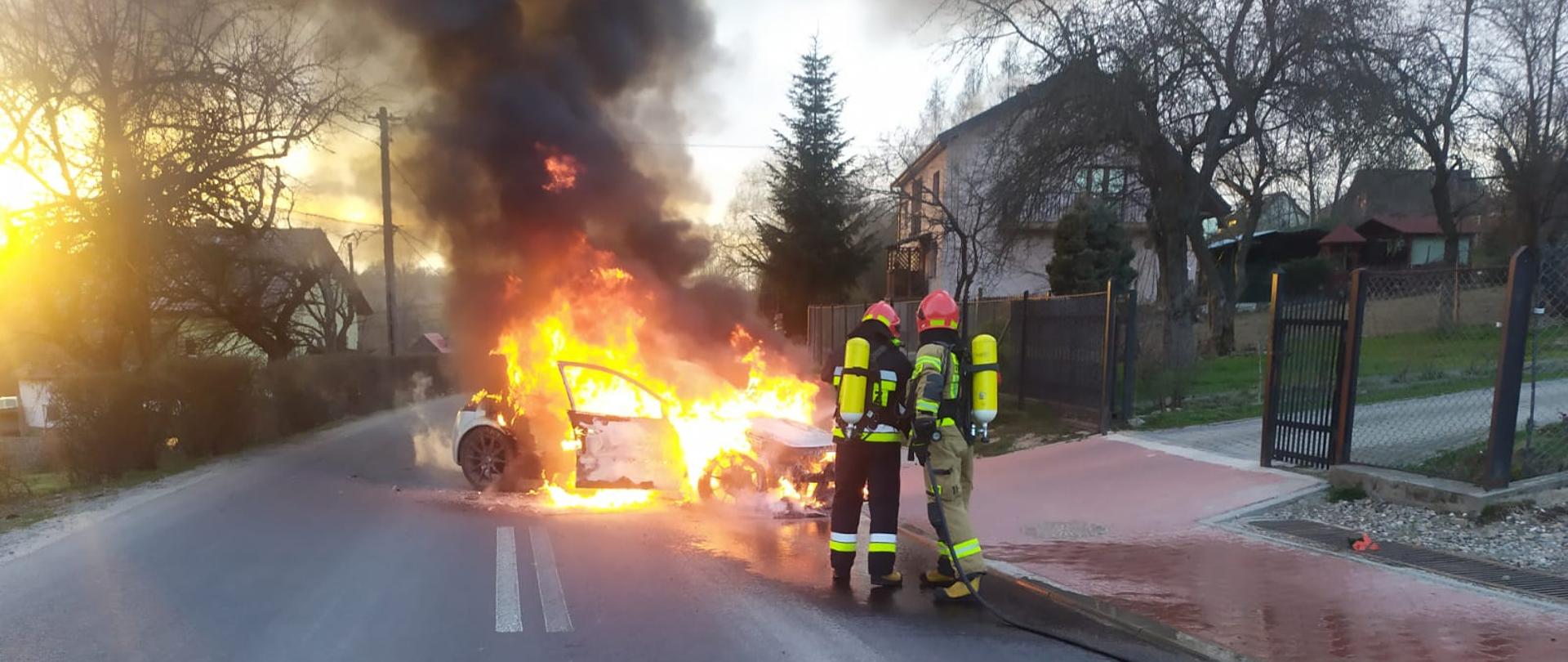 Na jezdni widoczny jest objęty ogniem samochód osobowy. Przed nim dwóch strażaków w umundurowaniu specjalnym z linią szybkiego natarcia podejmujących działania gaśnicze. Na drugim planie widoczne zabudowania miejscowości.