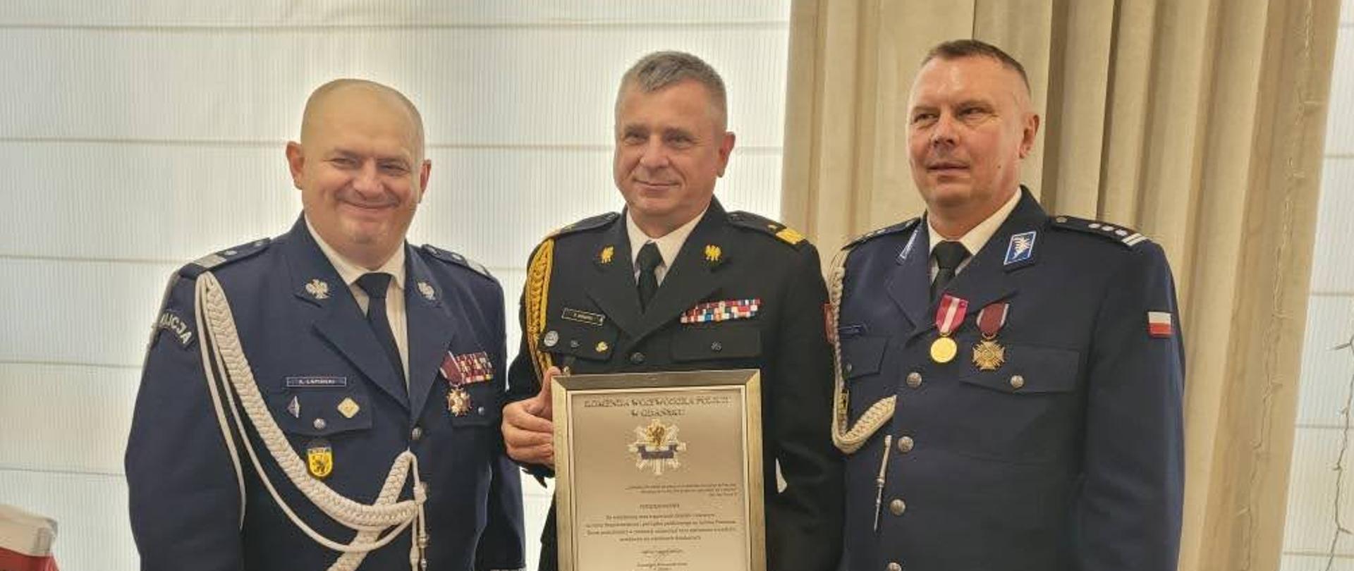 Pomorski komendant wojewódzki Państwowej Straży Pożarnej stoi trzymając wyróżnienie obok stoi kierownictwo Komendy Wojewódzkiej Policji.