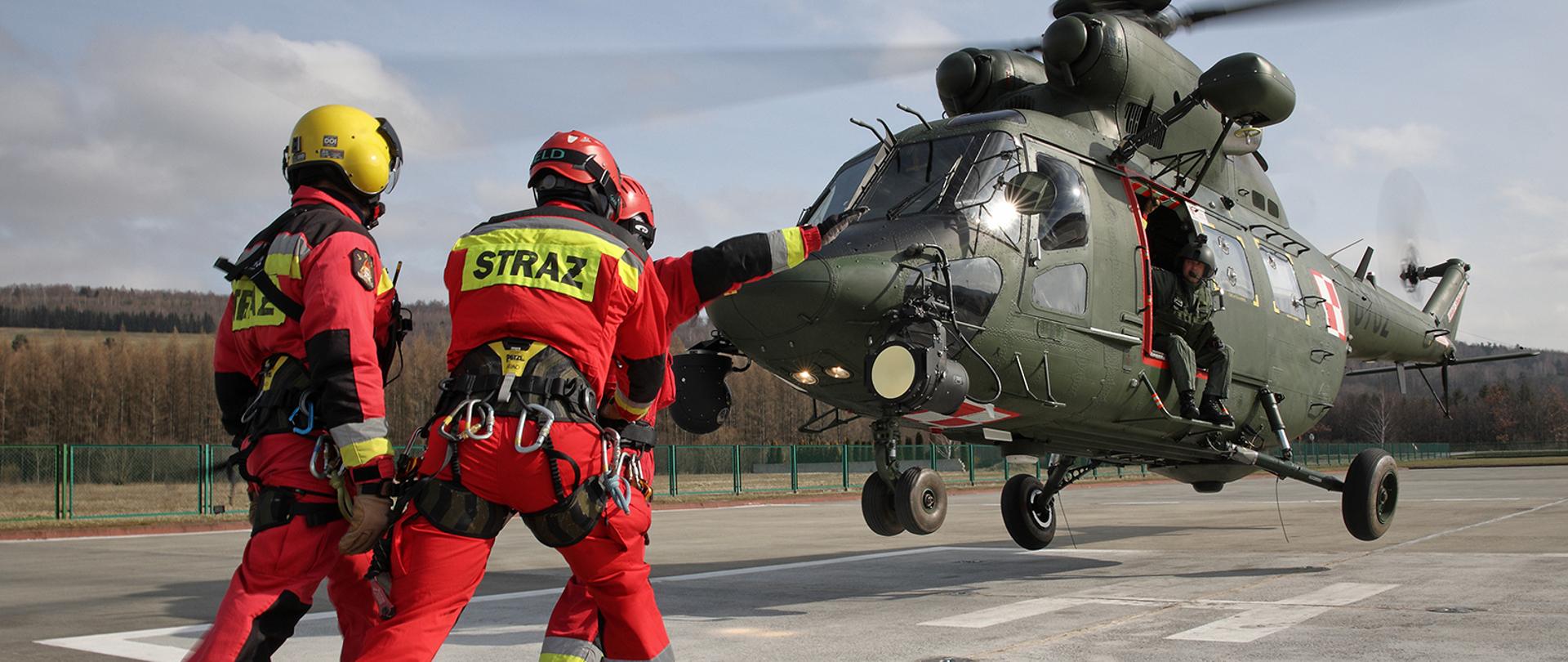 Zdjęcie przedstawia trzech strażaków wyposażonych w sprzęt ratownictwa wysokościowego oczekujących na wejście do lądującego helikoptera z którego wychylona jest jedna osoba.
