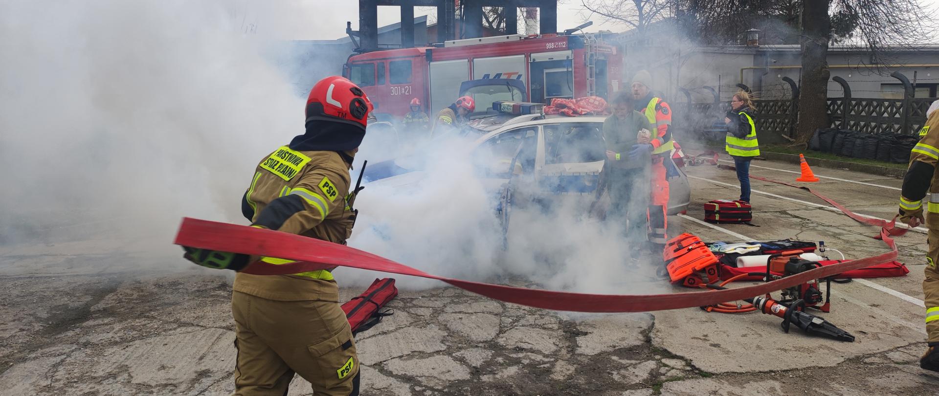 Symulacja wypadku z udziałem samochodu osobowego, zadymienie, na pierwszym planie strażaka rozwijający linie gaśniczą