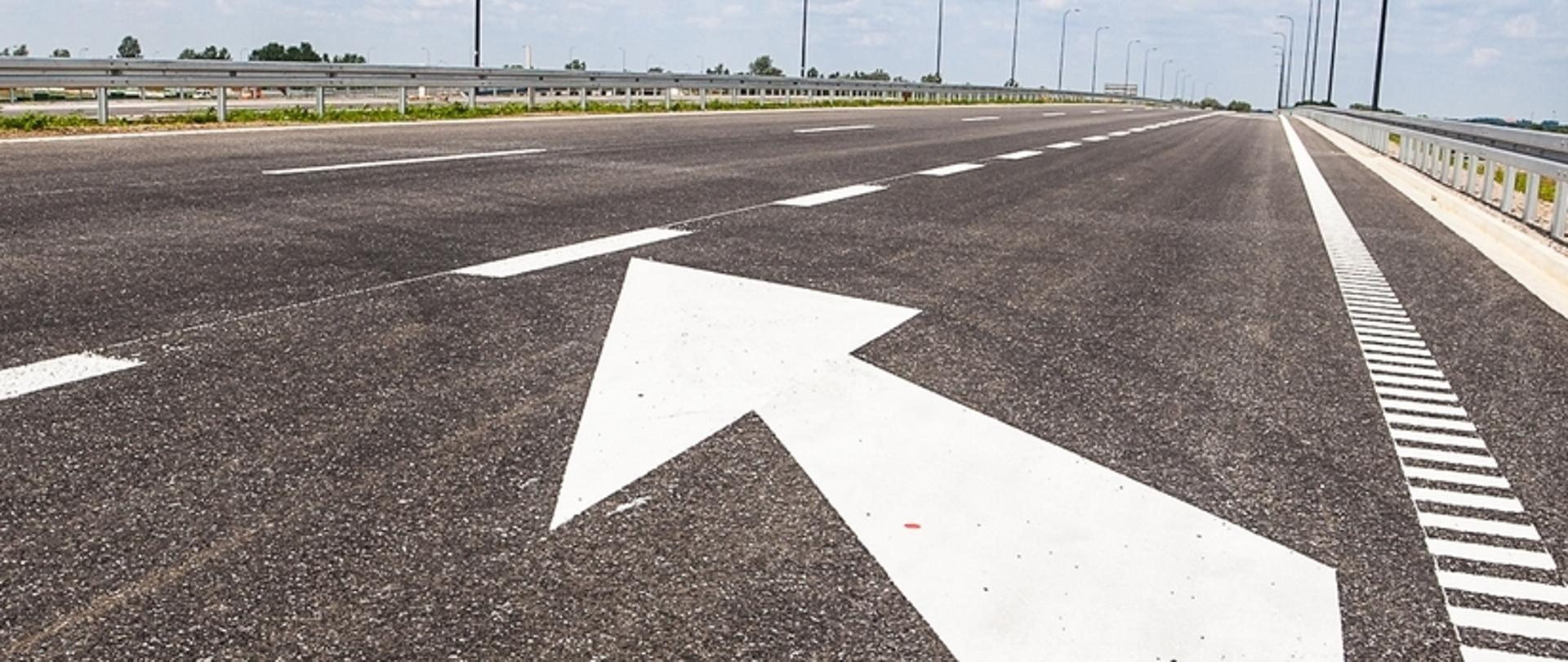 Zdjęcie przedstawia fragment autostrady A4. W centralnej części kadru widoczna jest szeroka jezdnia z oznakowaniem poziomym w postaci białej strzałki kierunkowej wskazującej jazdę na wprost. Po obu stronach drogi rozmieszczone są metalowe bariery ochronne, które zapewniają bezpieczeństwo użytkowników drogi. W tle można dostrzec dalszy odcinek autostrady, latarnie uliczne oraz otaczający krajobraz, prawdopodobnie składający się z pól lub łąk.