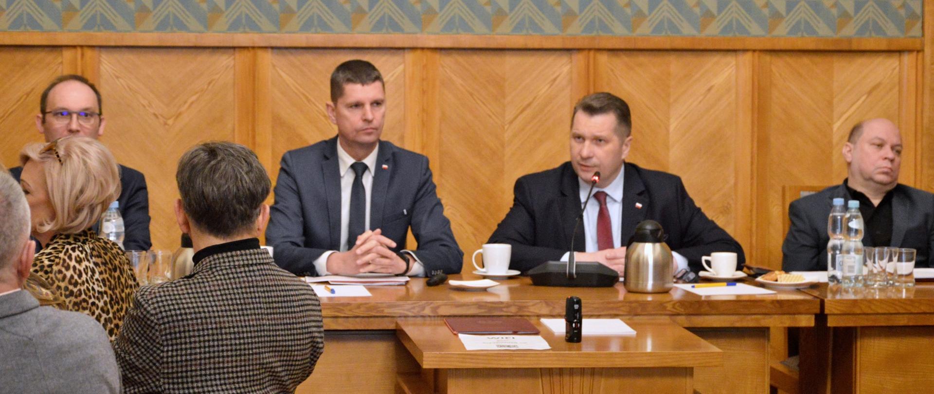 U szczytu drewnianego stołu siedzą minister Czarnek i wiceminister Piontkowski, minister mówi do małego mikrofonu, po obu stronach siedzą ludzie, za nimi zielone ściany częściowo wyłożone drewnem.