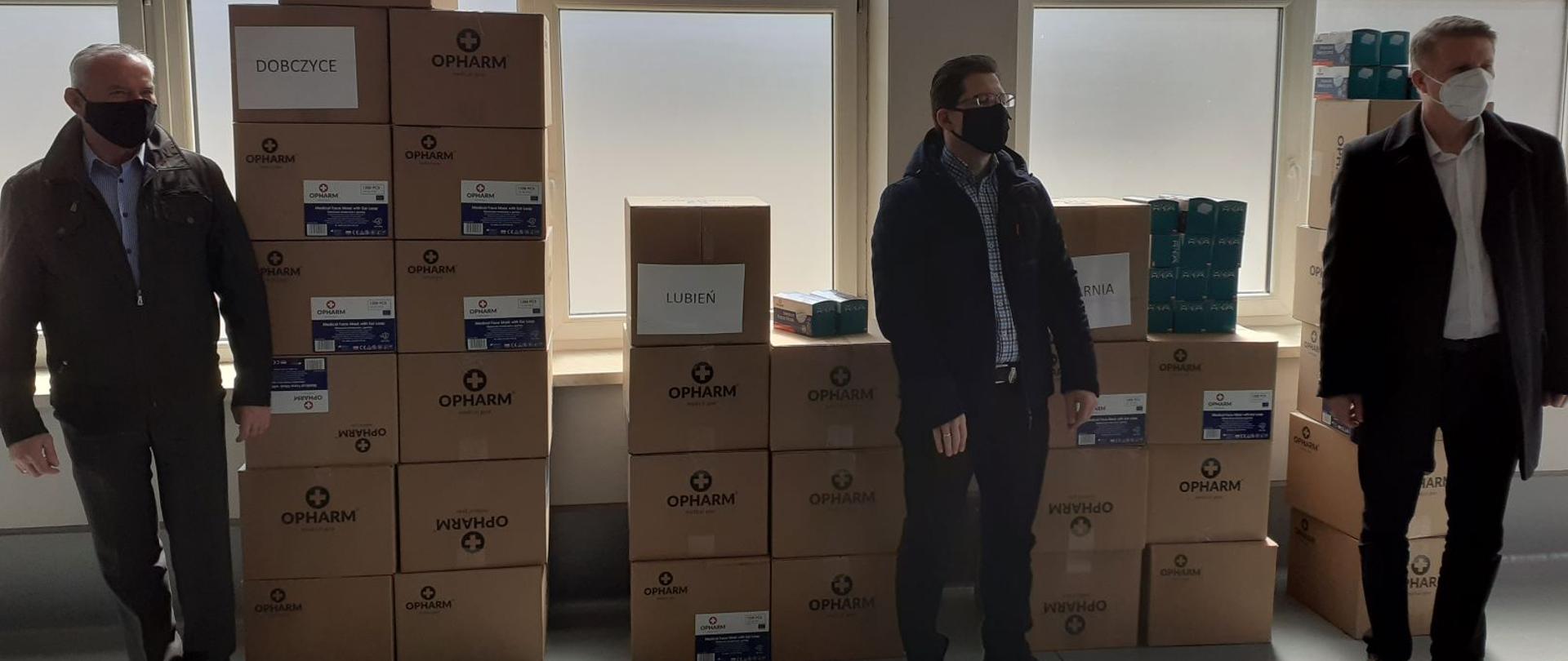 Na zdjęciu widzimy wnętrze garażu w Komendzie Powiatowej Państwowej Straży Pożarnej w Myślenicach, przygotowane pakiety maseczek ochronnych w pudłach tekturowych gotowe do odbioru przez przedstawicieli gmin. Przy pakietach stoją przedstawiciele z gmin, którzy przybyli aby je odebrać i przekazać mieszkańcom na swoim terenie. 