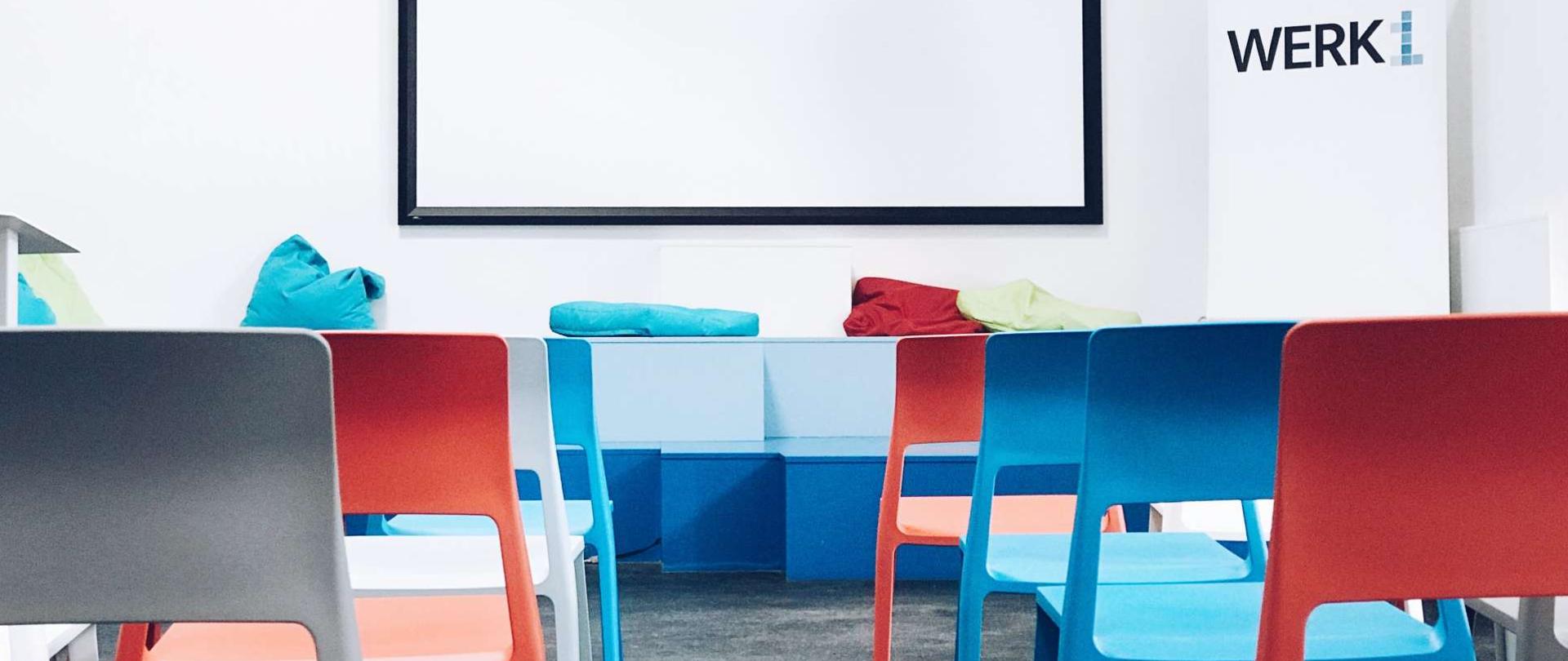 zdjęcie przedstawiające salę z białą tablicą i kolorowymi krzesłami ustawionymi w dwóch rzędach