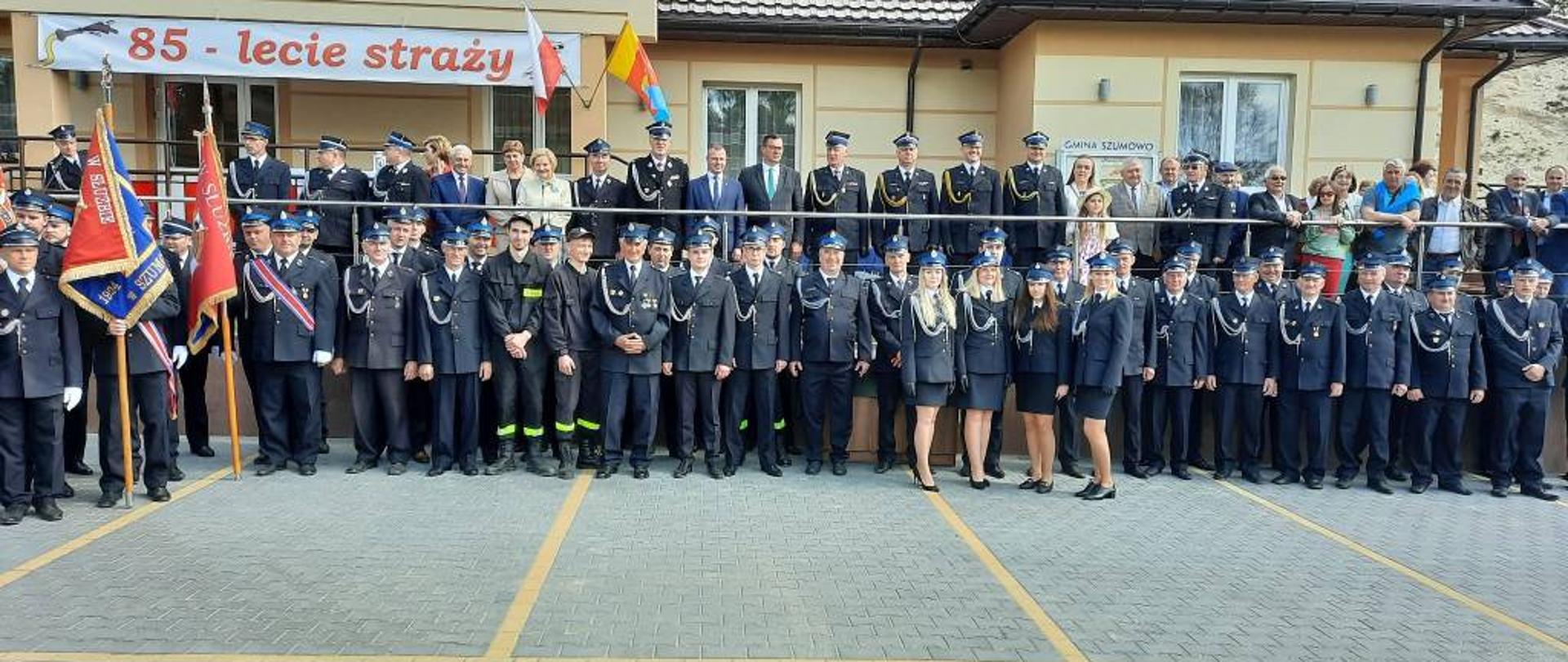Zdjęcie przedstawia wszystkich uczestników spotkania okolicznościowego z okazji 85-lecia powstania Ochotniczej Straży Pożarnej w Zarębach-Jartuzach pozujących do zdjęcia na tle budynku remizy strażackiej. Większość uczestników ubrana w mundury wyjściowe Ochotniczej Straży Pożarnej ze sznurem galowym. Poza strażakami na zdjęciu znajdują się również inni uczestnicy spotkania w ubraniach cywilnych. Zdjęcie wykonano w słoneczny dzień.