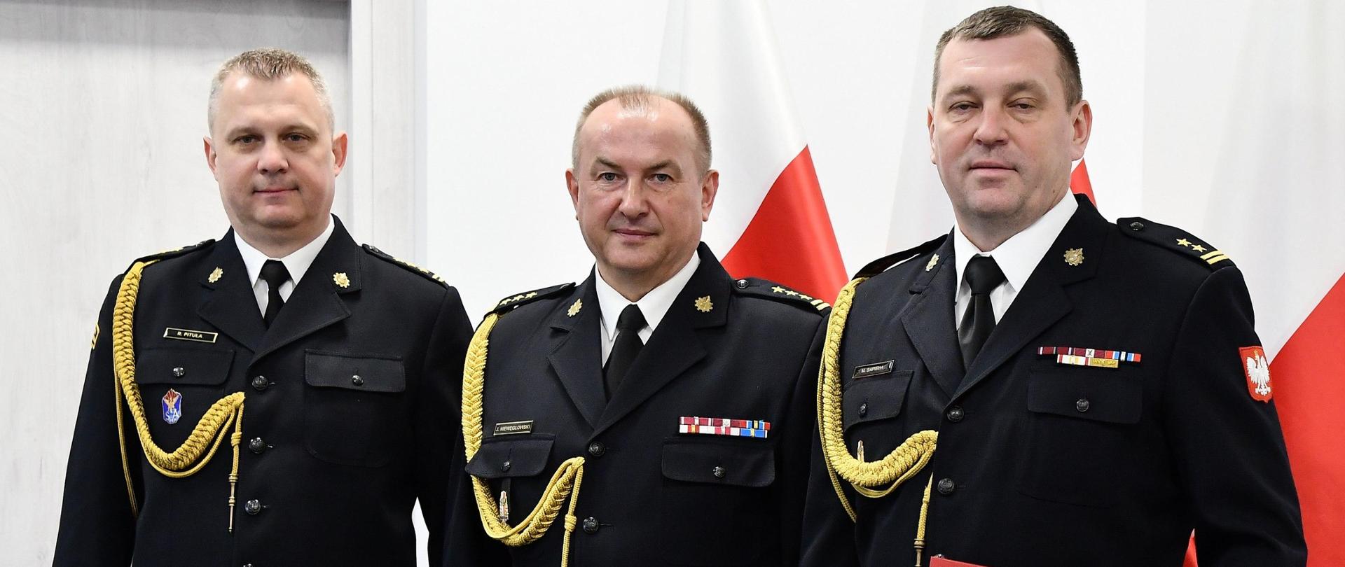 Trzech strażaków w mundurach wyjściowych ze sznurem stoi obok siebie jeden z mężczyzn trzyma czerwoną teczkę za nimi ustawione są trzy flagi Polski oraz znajdują się drzwi. 