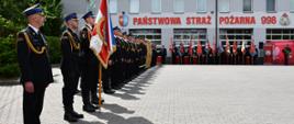 Pododdział strażaków podczas apelu na placu przed budynkiem Komendy Powiatowej Państwowej Straży Pożarnej, na którym widnieje duży, czerwony napis „PAŃSTWOWA STRAŻ POŻARNA 998”. Po lewej stronie na pierwszym planie stoją Dowódca uroczystości oraz poczet sztandarowy. Dalej za nimi oraz wzdłuż budynku, w równych szeregach stoi kilkudziesięciu strażaków.