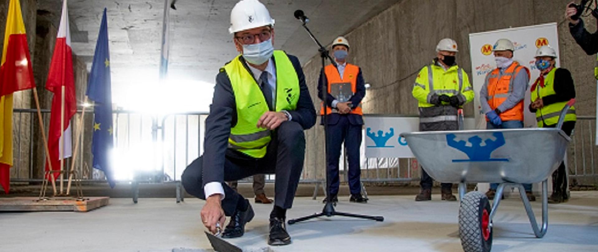 Zdjęcie przedstawia ministra umieszczającego kamień węgielny w podziemiach metra