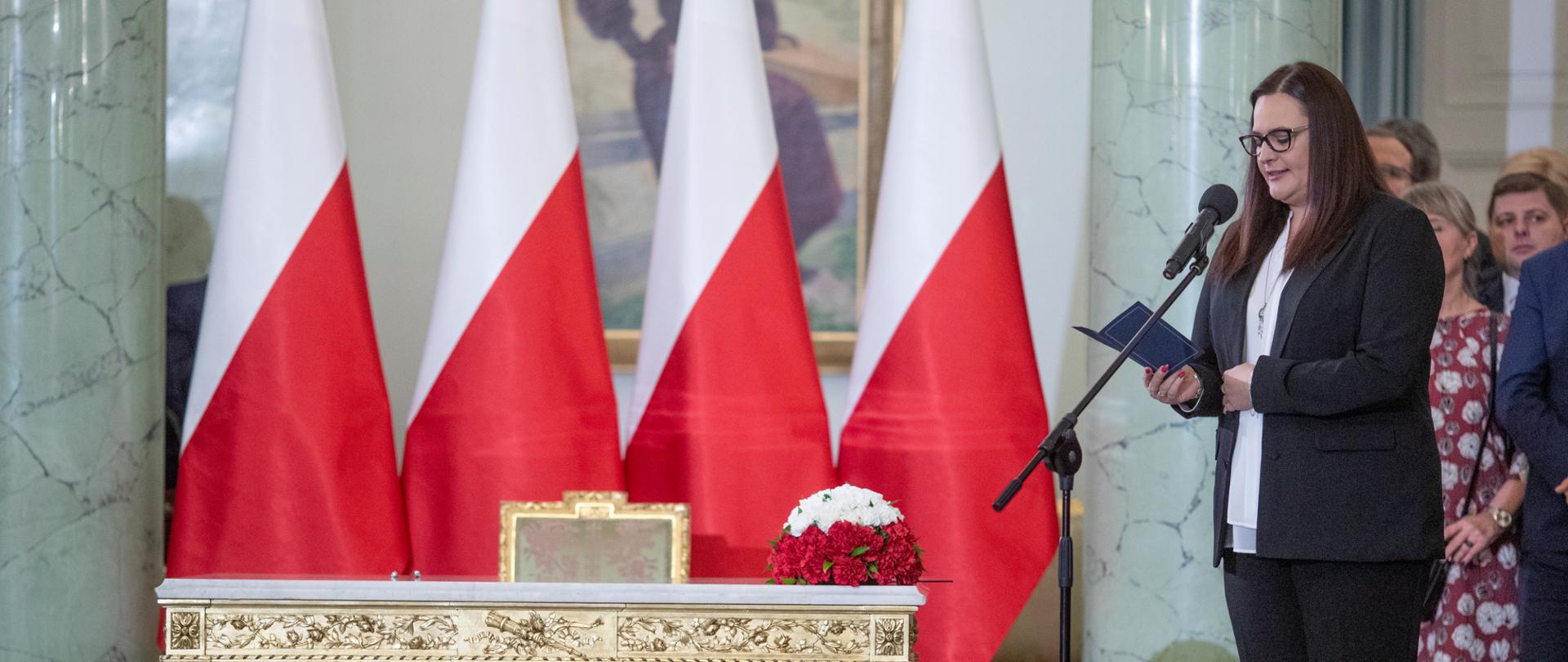 Minister odczytuje tekst roty przysięgi, stojąc przy mikrofonie. Na środku biało-czerwone flagi RP