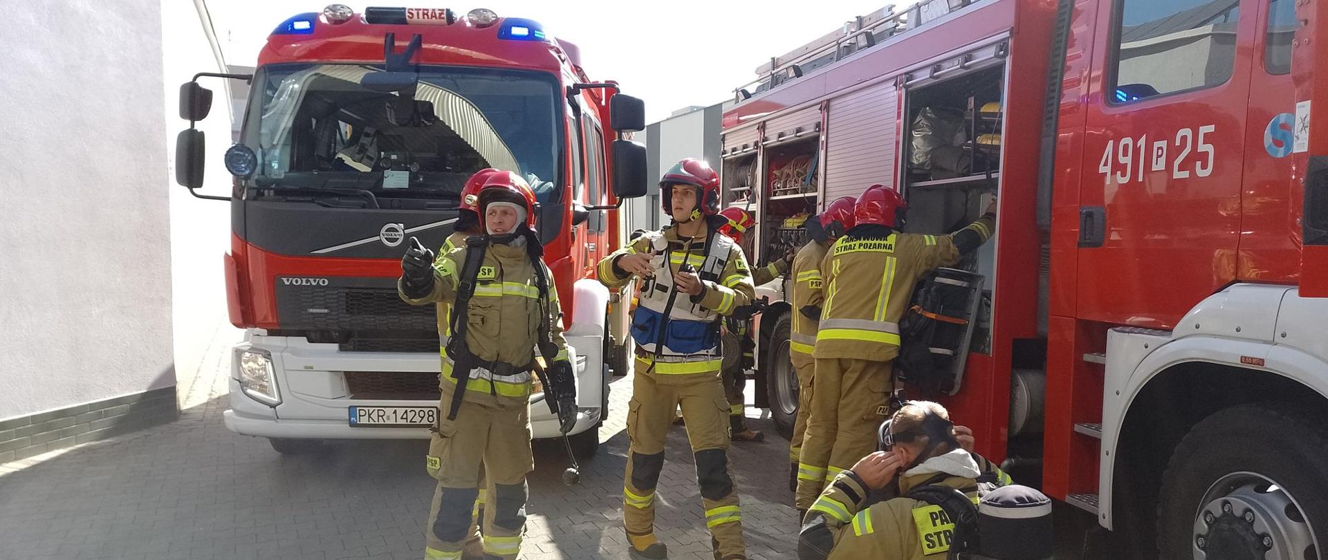 Zdjęcie przedstawia strażaków przy wozach strażackich, podczas przygotowywania się do wejścia w strefę niebezpieczną.