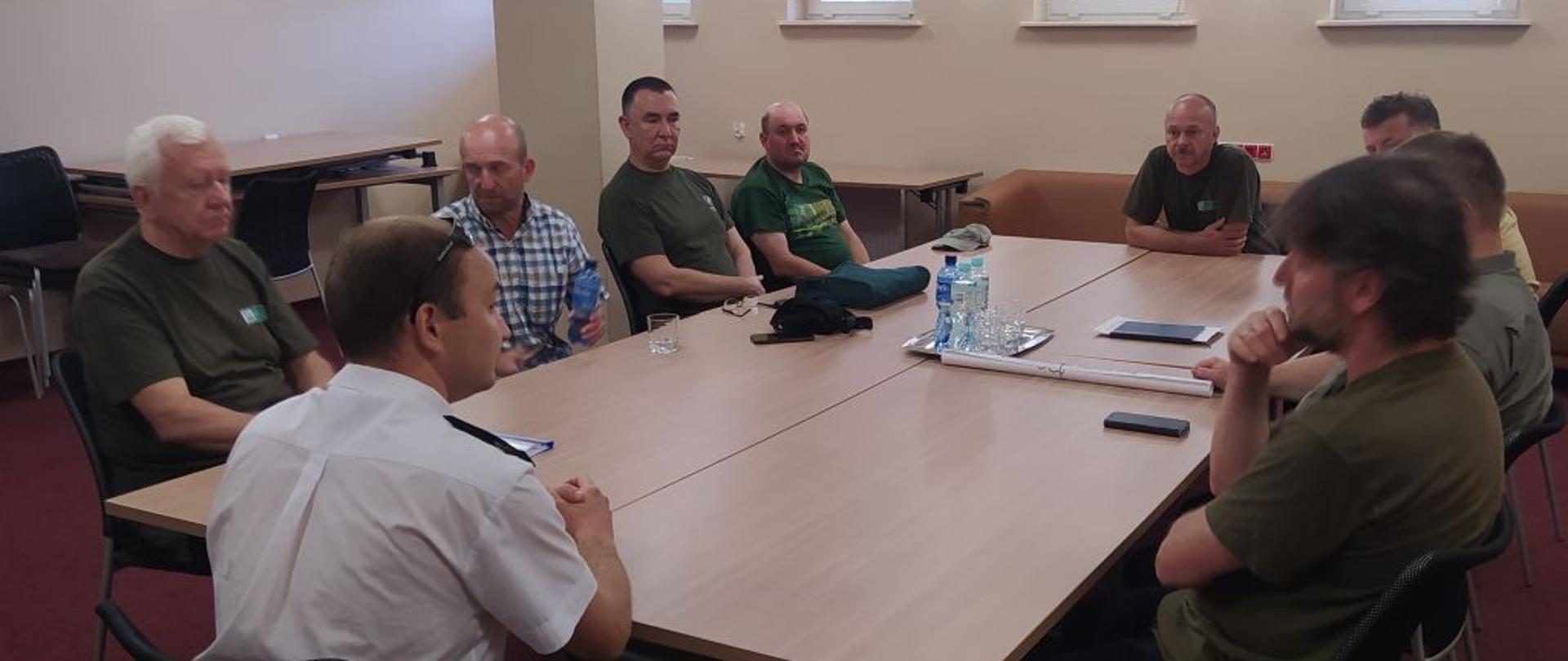 Przy prostokątnym stole siedzi dziewięciu mężczyzn w zielonych koszulkach. Odwrócony plecami siedzi mężczyzna w białej koszuli.