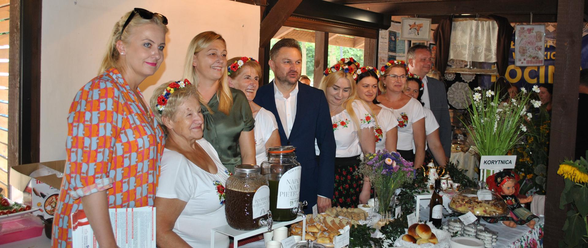 Grupa osób w tym członkinie KGW w wiankach z kwiatów i zaproszeni goście pozuje do zdjęcia za stołem zastawionym potrawami i napojami