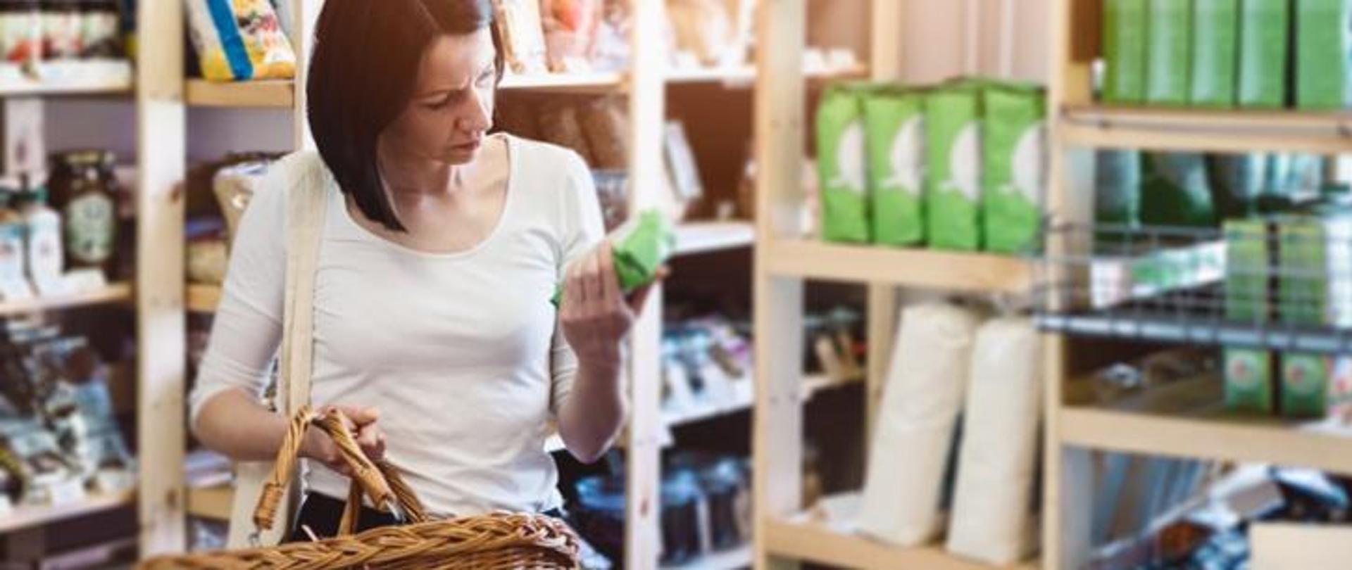 Kobieta w sklepie spożywczym, trzyma w prawej ręce koszyk na zakupy a w lewej dłoni produkt spożywczy i czyta etykietę