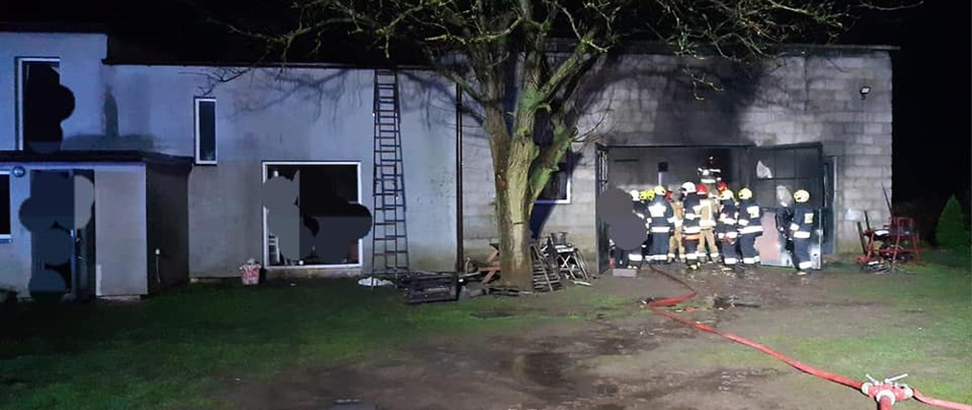 Na zdjęciu widać budynek murowany parterowy, w którym doszło do pożaru. Do pożaru doszło w pomieszczaniu w prawej części budynku. W wejściu do garażu stoi grupa kilkudziesięciu strażaków. Przed budynkiem rośnie drzewo. Na ziemi widać czerwony wąż strażacki z rozdzielaczem. Jest ciemno, ale miejsce jest oświetlone.