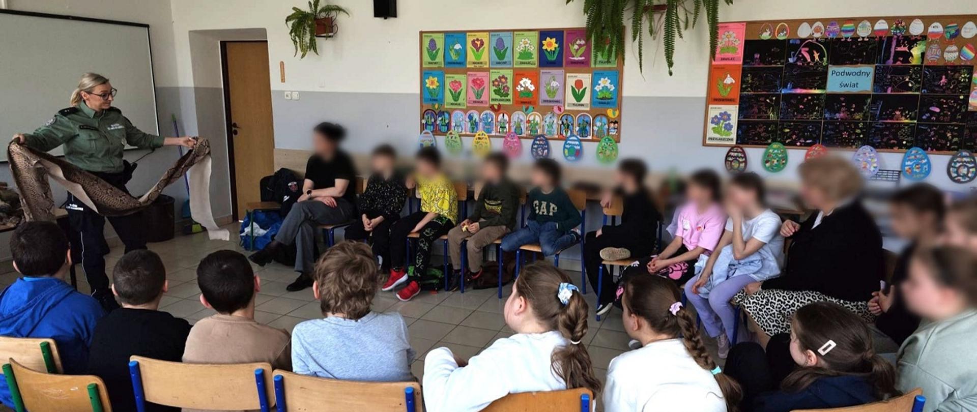 Sala lekcyjna. Uczniowie siedzą na krzesłach ułożonych w koło. Funkcjonariuszka SCS trzyma w dłoniach skórę chronionego gada.. 