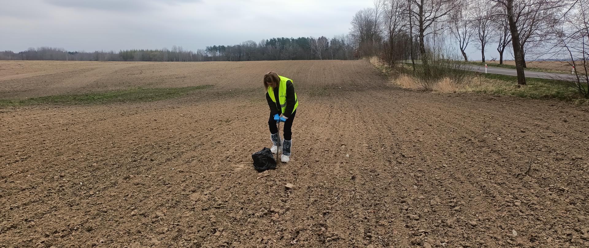 Kobieta na uprawionym polu w ochraniaczach na buty, rękawiczkach i kamizelce odblaskowej, trzyma próbobierz i pobiera próby