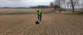 Kobieta na uprawionym polu w ochraniaczach na buty, rękawiczkach i kamizelce odblaskowej, trzyma próbobierz i pobiera próby