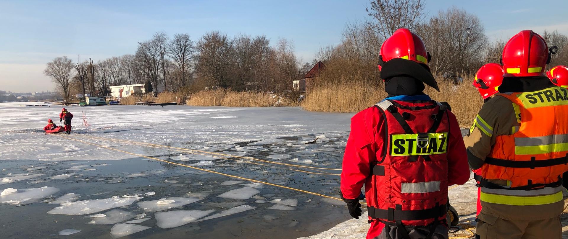 Zamarznięte jezioro. Na brzegu grupa strażaków - ćwiczenia- wyciąganie po linie poszkodowane. Wokół wysokie trawy, zabudowania.
