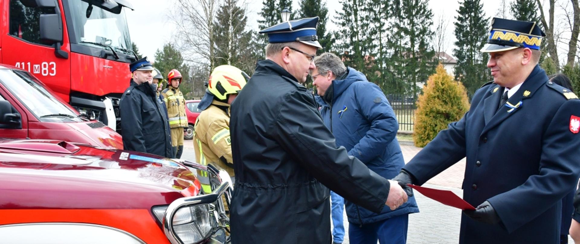 Na zdjęciu widoczni minister Maciej Wąsik i mazowiecki komendant wojewódzki PSP nadbrygadier Jarosław Nowosielski w trakcie wręczania dokumentów strażakom ochotnikom stojącym przy samochodzie strażackim.