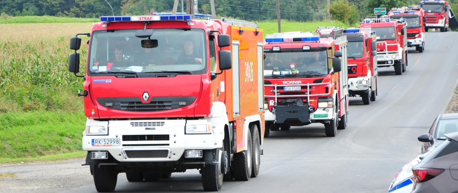 Zdjęcie przedstawia jadącą po drodze publicznej kolumnę ciężkich samochodów pożarniczych